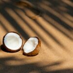 Coconut halves resting on a sunlit sandy beach, casting shadows from palm fronds.